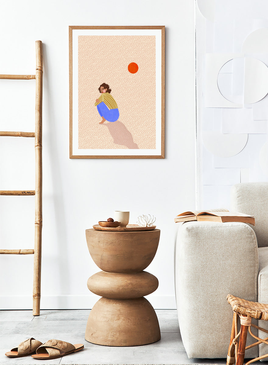 A minimalist art print featuring a stylized illustration of a person crouching in blue pants and a yellow striped top, looking up at a small orange sun against a light beige dotted background, framed with a white mat and black border. Pensive Woman Under Sun by Bea Müller art print featuring a seated woman beneath an orange sun in bold blue, mustard, and soft neutral tones.