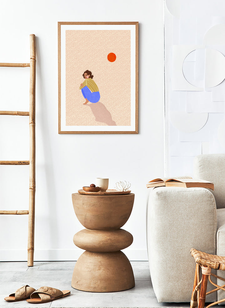 A minimalist art print featuring a stylized illustration of a person crouching in blue pants and a yellow striped top, looking up at a small orange sun against a light beige dotted background, framed with a white mat and black border. Pensive Woman Under Sun by Bea Müller art print featuring a seated woman beneath an orange sun in bold blue, mustard, and soft neutral tones.