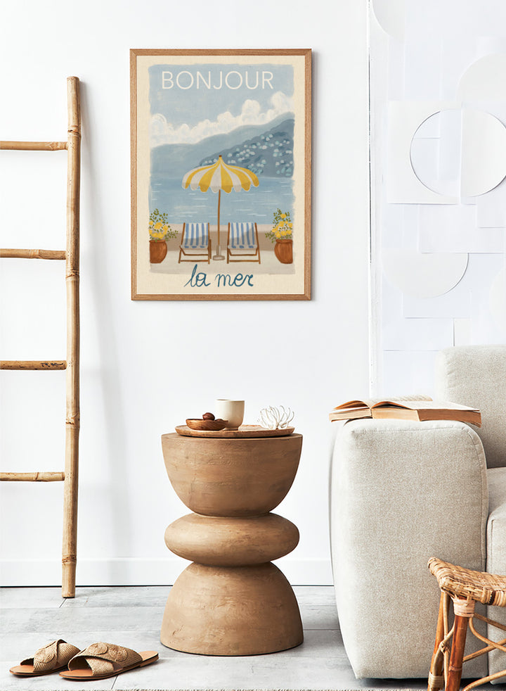 A vintage-style illustrated poster or print showing a beach scene with two striped lounge chairs under a yellow-and-white umbrella, potted plants, blue water, and mountains in the background. The words &