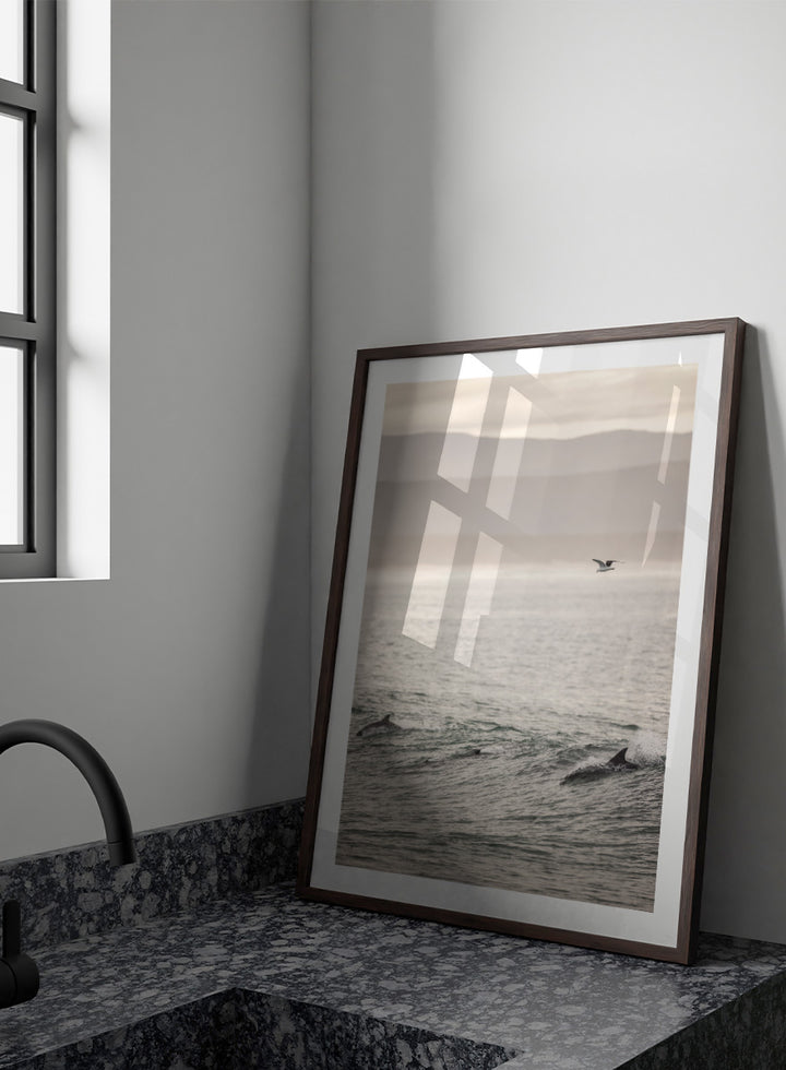 A framed photographic art print depicting dolphins swimming in a misty ocean with a bird flying in the distance. Ocean’s Dance by Shot by Clint ocean photography print featuring dolphins, rolling waves, a flying bird, and hazy coastal hills.