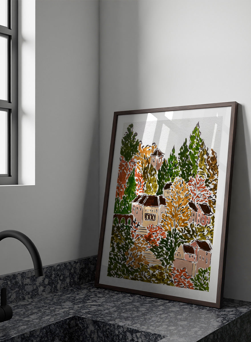 A framed art print featuring a hand-drawn, stylized landscape of a hillside village with beige buildings, brown roofs, and dense trees in green, yellow, orange, and red tones. Shop Autumn Hillside Village by Rushati Chowdhury, a colorful autumn landscape art print with hillside homes, winding steps, dense trees, and warm fall tones.