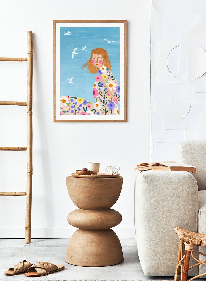 A framed art print featuring a stylized illustration of a young woman with long light brown hair, surrounded by colorful flowers and white birds against a light blue sky. ree Spirit Floral Dreams by Bea Müller floral woman art print featuring a flowing flower-covered summer dress, white birds, and a bright blue sky.
