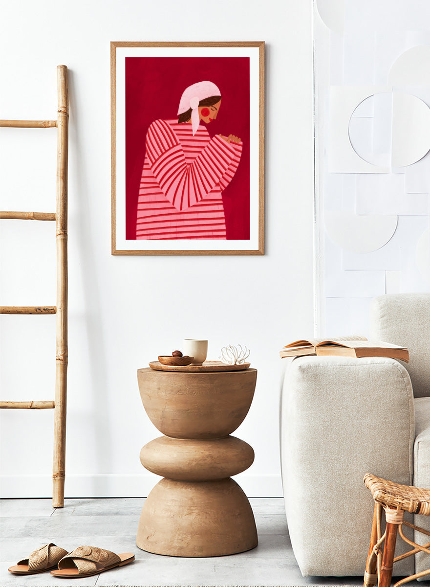 A framed art print featuring a stylized illustration of a woman in a pink and red striped top and white headscarf against a deep red background. Serene Praying Woman by Bea Müller portrait art print featuring a reflective woman in a pink striped dress against a rich red background.