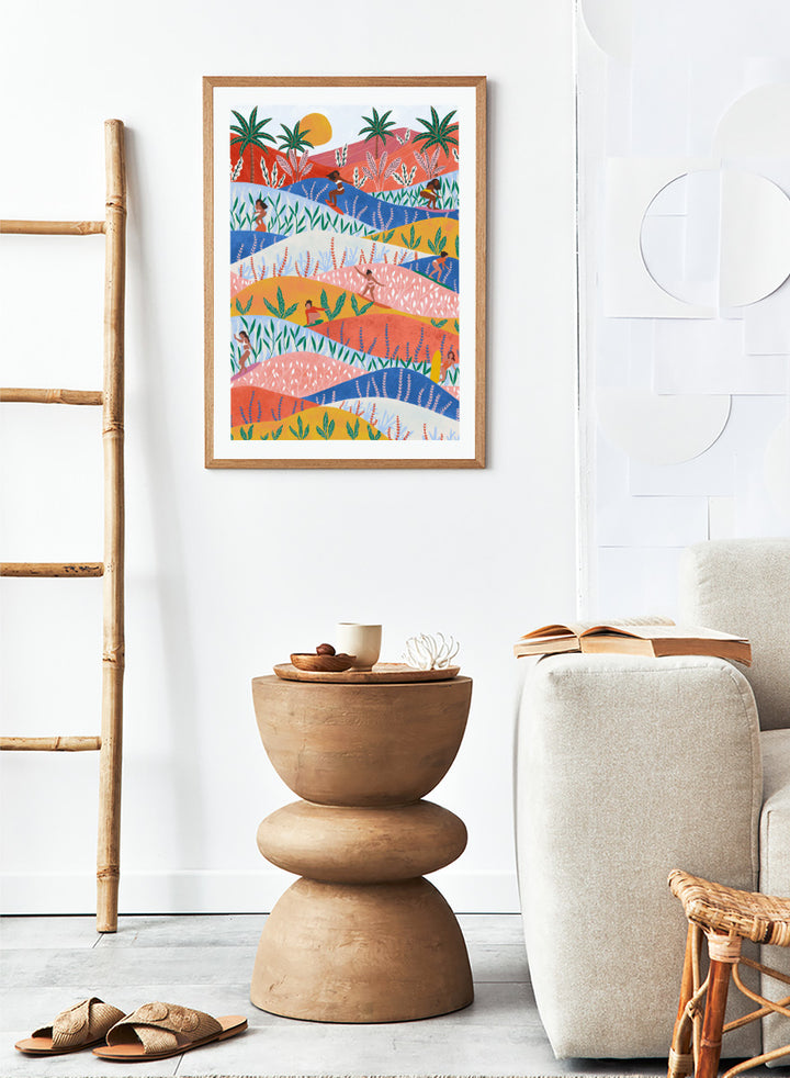 A framed art print featuring a colorful, stylized landscape illustration with rolling hills, palm trees, a sun, and small human figures in a folk-art style. Tropical Surf Girls by Ceyda Alasar surf art print featuring women riding waves through a colorful palm-filled landscape in bold tropical tones.