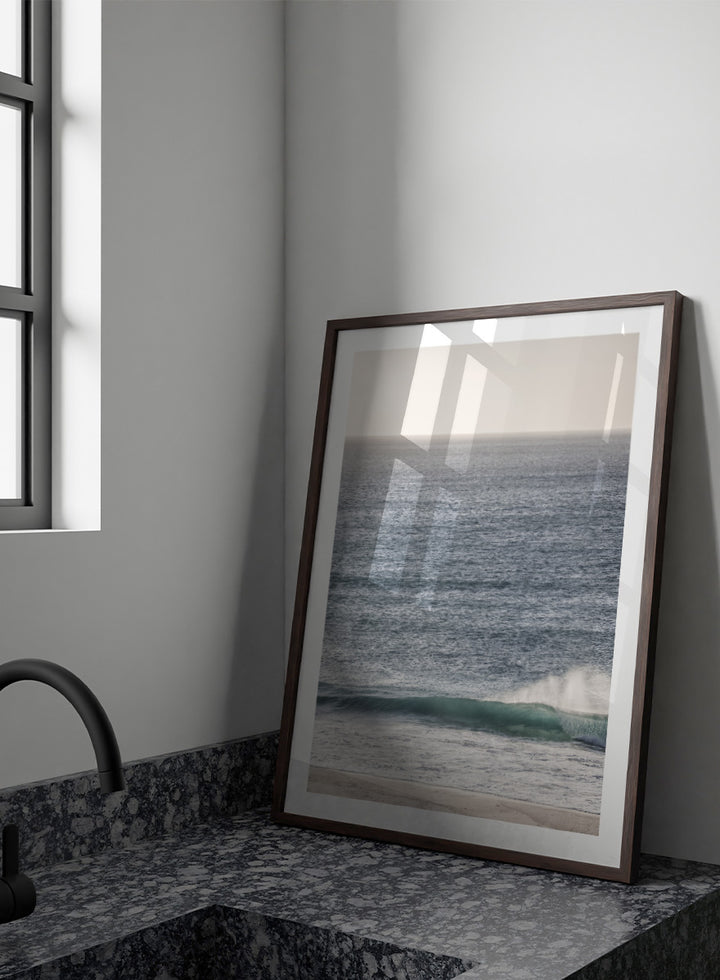 A framed photographic art print of an ocean scene, featuring a gentle wave breaking on a sandy beach under a pale sky. Tranquil Ocean Scene by Shot by Clint ocean photography print with a rolling wave, soft sea spray, and airy blue coastal tones.