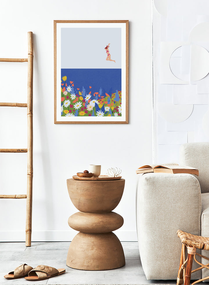A framed art print featuring a minimalist illustration of a woman in a pink bikini jumping into a blue sea with a colorful wildflower meadow below. Wildflower Summer Jump art print by Ceyda Alasar features a joyful figure above colorful blooms, bringing playful summer energy to your walls.