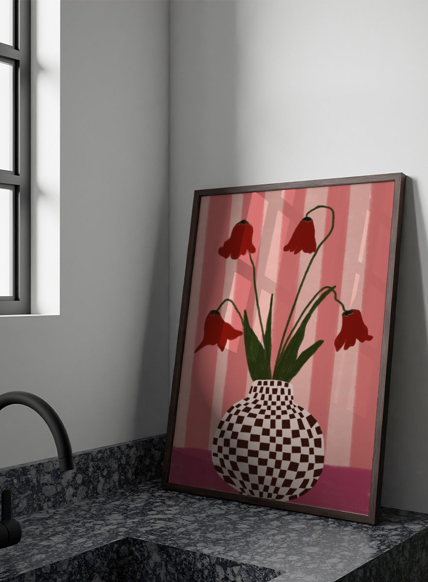 A stylized floral artwork showing four dark red bell-shaped flowers with long green stems rising from a black-and-white checkerboard vase, set against pink vertical stripes and a pink base. Whimsical Flowers Checkered Vase by The Art Concept still life art print featuring red flowers, a checkered vase, and a soft pink striped background.