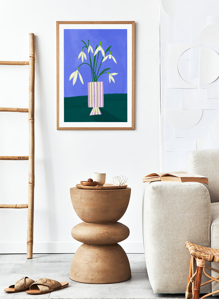 A framed art print featuring a stylized illustration of white snowdrops with green stems in a lavender and cream striped vase, set against a blue and dark green background. Snowdrops in Striped Vase by Bea Müller fine art print featuring white snowdrops, a lilac striped vase, and a deep blue background.