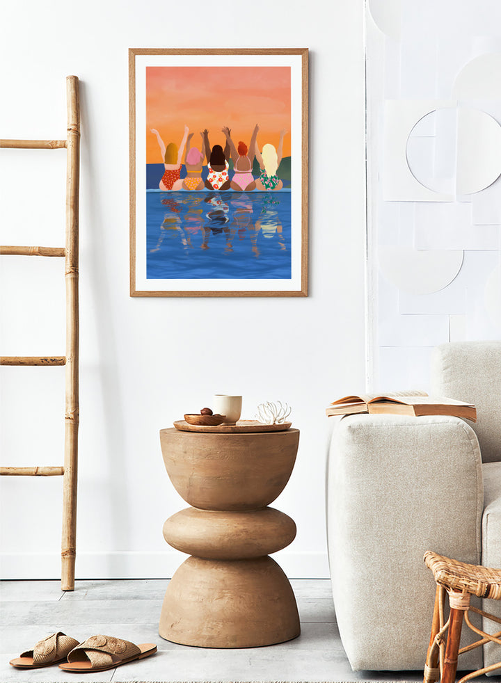 A framed art print featuring a stylized illustration of five women sitting in a pool at sunset, seen from behind as they raise their arms. The background is a warm orange sky, and the water below reflects their silhouettes. Summer Sunset Friends by Ceyda Alasar friendship art print featuring five women by the water in patterned swimsuits beneath a glowing orange sky.