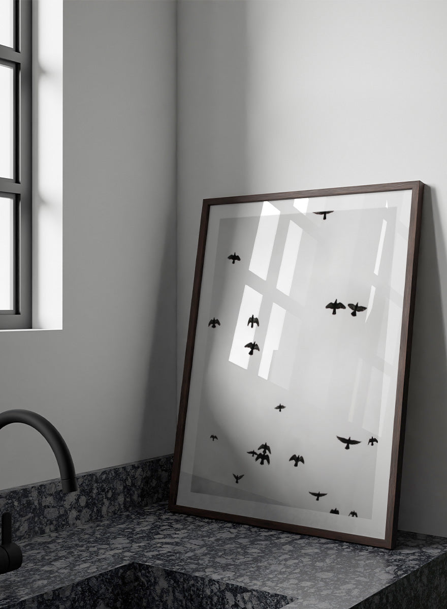 A framed black and white art print depicting a flock of birds flying across a cloudy sky, presented in a minimalist look. Birds in Flight by Shot by Clint black-and-white bird photography print featuring a scattered flock, open sky, and graphic silhouettes in motion.