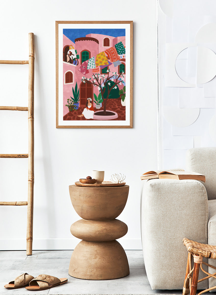 A framed art print featuring a colorful Mediterranean-style illustration of a pink courtyard with a woman sitting under a flowering tree, laundry hanging in the background, and a blue sky above. Pink Village Courtyard by Ceyda Alasar art print featuring pink architecture, patterned rugs, flowering trees, potted plants, and relaxed courtyard life.