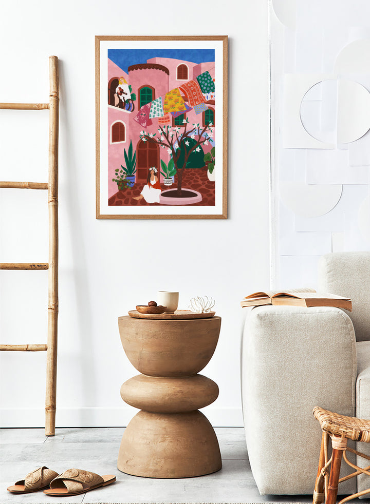 A framed art print featuring a colorful Mediterranean-style illustration of a pink courtyard with a woman sitting under a flowering tree, laundry hanging in the background, and a blue sky above. Pink Village Courtyard by Ceyda Alasar art print featuring pink architecture, patterned rugs, flowering trees, potted plants, and relaxed courtyard life.