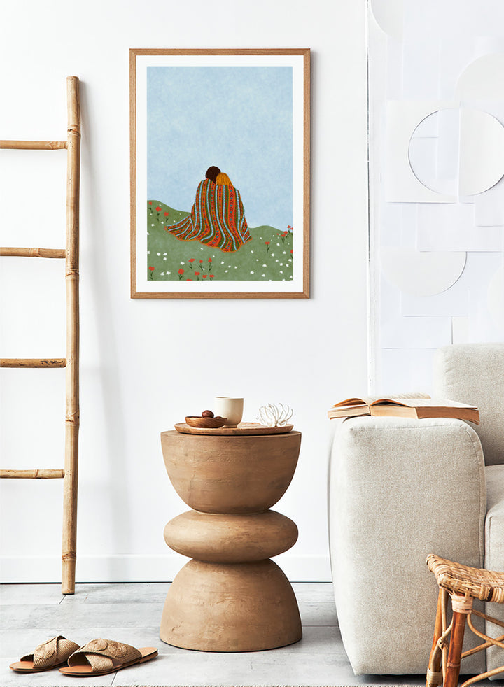 A framed art print featuring an illustration of two people sitting on a grassy hill, wrapped in a vibrant, patterned blanket under a light blue sky. Cozy Hillside Embrace by Ceyda Alasar art print featuring two people wrapped in a patterned blanket among green hills and wildflowers.