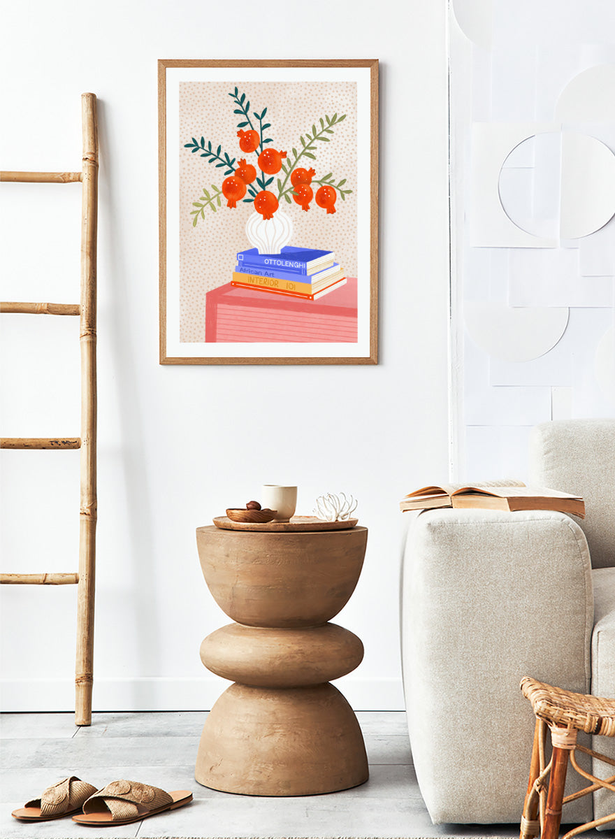 A framed art print featuring a stylized illustration of pomegranate branches in a white vase resting on a stack of books, set against a light beige dotted background. Cozy Tabletop Pomegranates by Bea Müller still life art print featuring red fruit branches, a sculptural vase, design books, and colorful interior styling.
