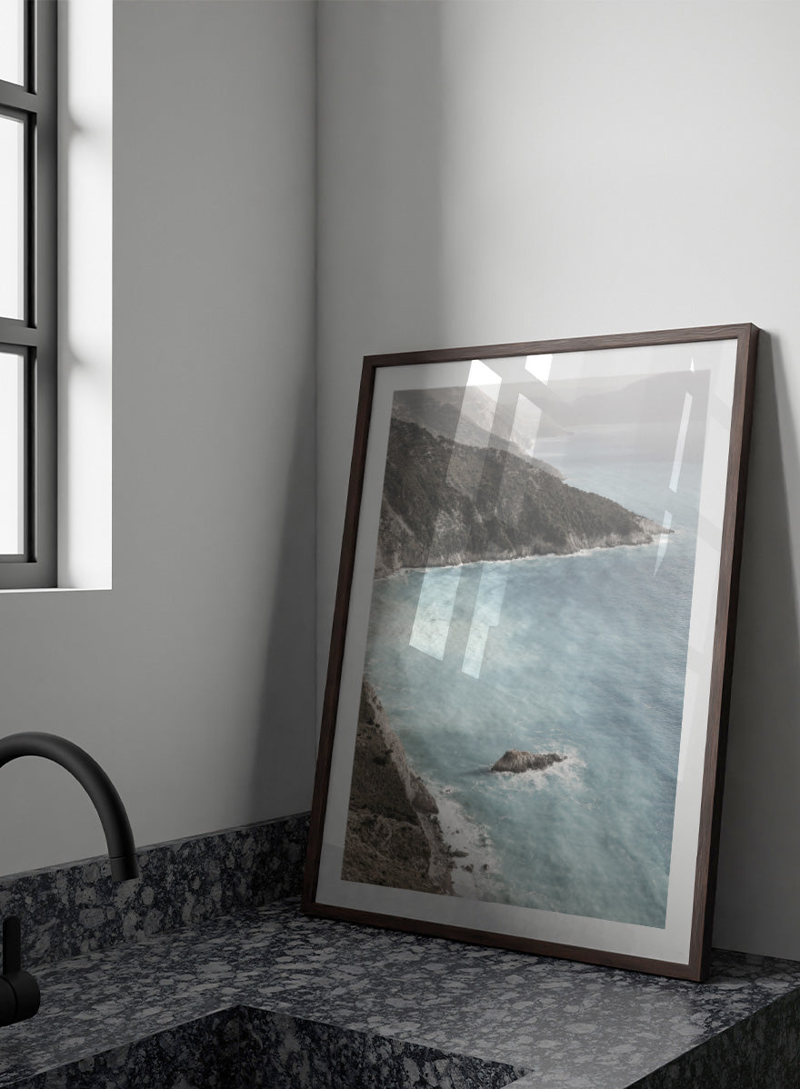 A framed photographic art print depicting a coastal landscape with a steep rocky cliff overlooking a calm, pale blue sea with a small rock formation in the foreground. Coastal Serenity by Shot by Clint coastal photography print featuring pale blue water, rugged cliffs, rocky shores, and a soft hazy horizon.