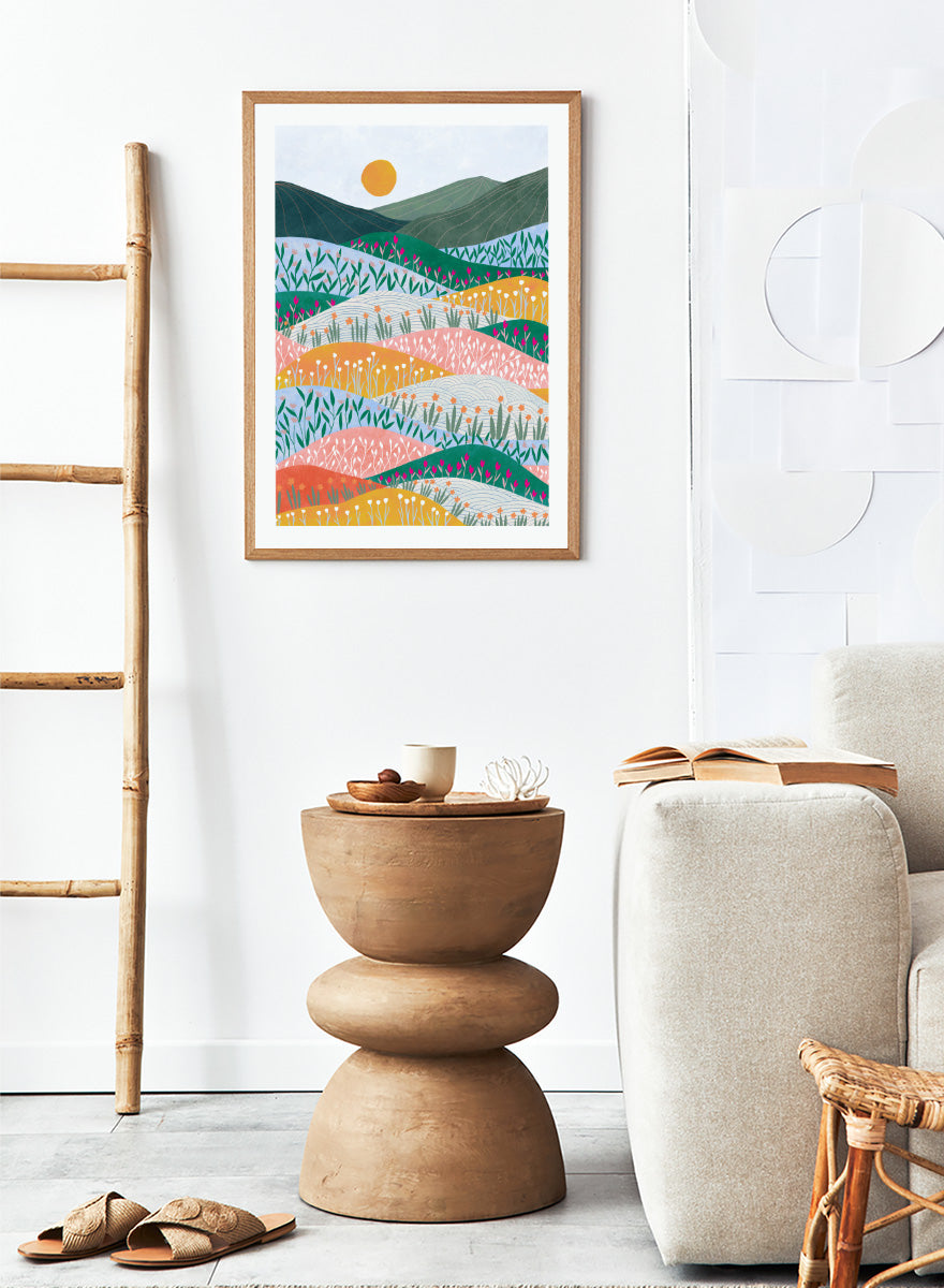 A framed art print featuring a stylized landscape illustration with rolling hills, wildflowers, mountains, and a sun in pastel and bright colors. Colorful Flower Hills by Ceyda Alasar botanical landscape art print featuring layered wildflower fields, rolling hills, mountains, and a golden sun.