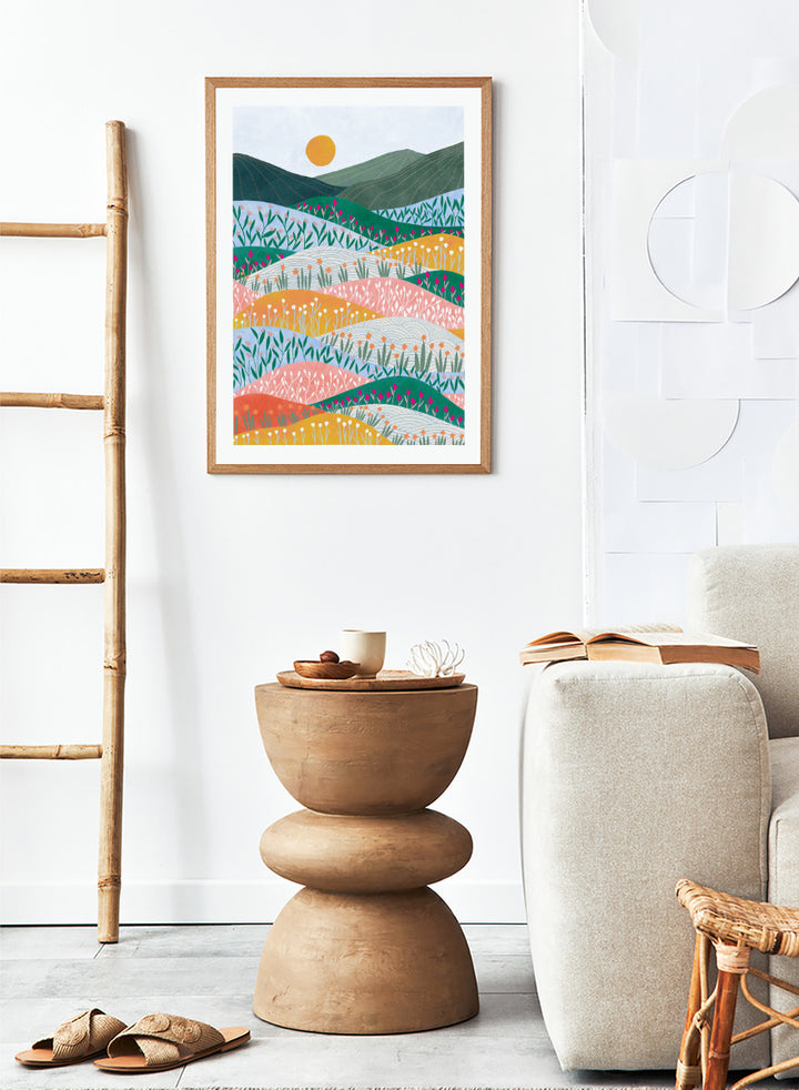 A framed art print featuring a stylized landscape illustration with rolling hills, wildflowers, mountains, and a sun in pastel and bright colors. Colorful Flower Hills by Ceyda Alasar botanical landscape art print featuring layered wildflower fields, rolling hills, mountains, and a golden sun.