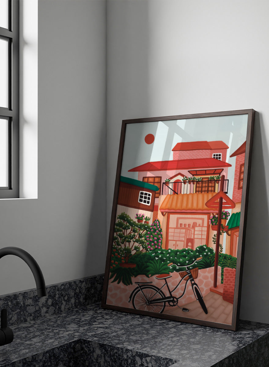A colorful illustrated street scene with pink and peach buildings, green plants, flowers, a bicycle in the foreground, and a pale sky with a red sun. Shop Charming Village Street by Nidhi Kachhadiya, a colorful village wall art print featuring a bicycle, flower-filled homes, terracotta roofs, and warm pink and green tones.
