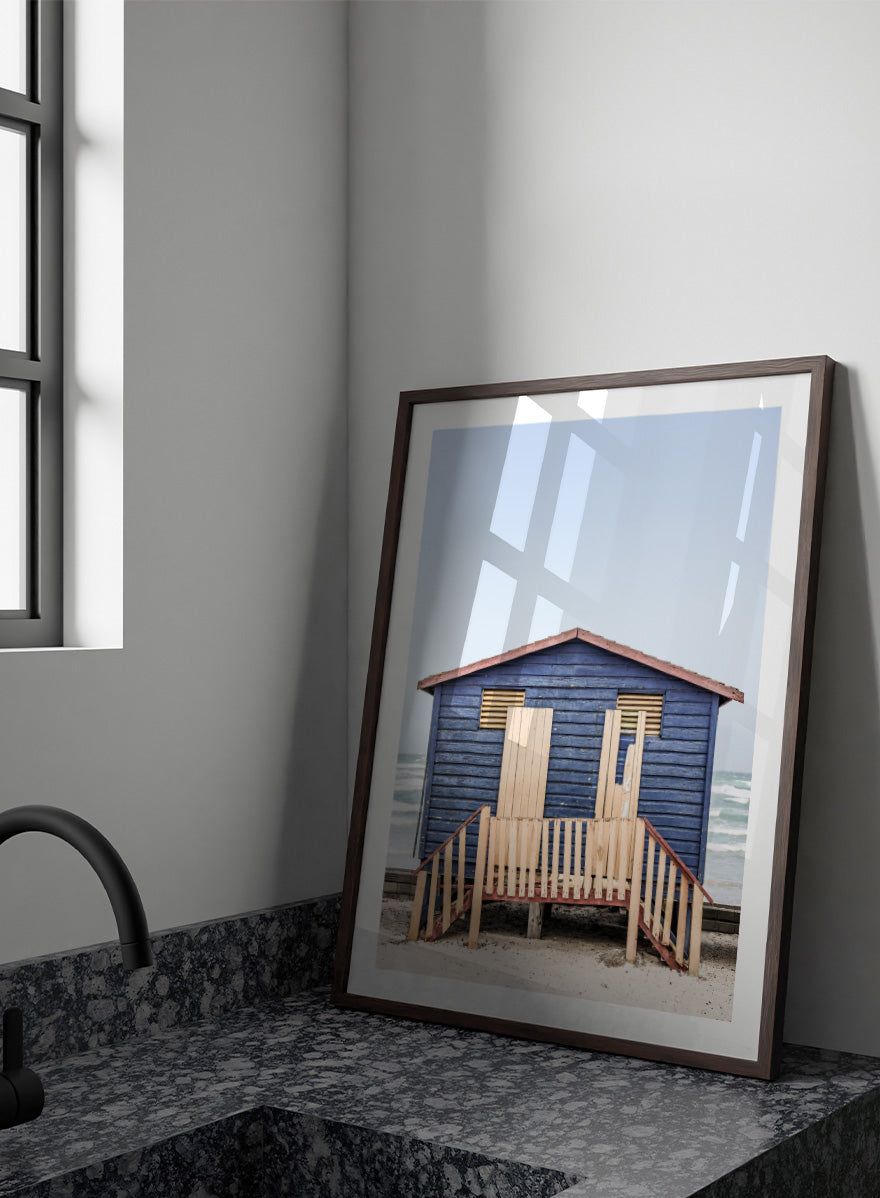 A framed photographic print of a blue wooden beach lifeguard hut on a sandy shore with the ocean and pale sky in the background. Beachside Retreat by Shot by Clint features a weathered blue beach hut and soft ocean backdrop in a calm coastal photography print.