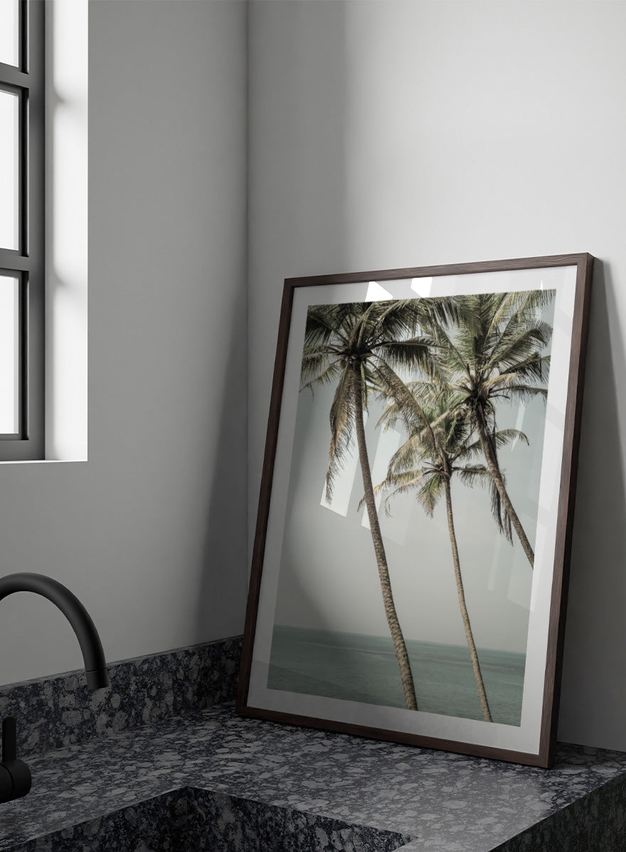 A framed art print featuring three leaning palm trees against a pale sky and ocean horizon. Ocean Breeze Palms by Shot by Clint coastal art print featuring tall palm trees, soft blue ocean views, and an airy tropical landscape.