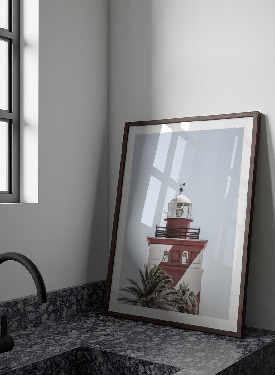 A framed art print featuring a red and white lighthouse with palm trees at the bottom, set against a pale sky background. Beacon of Solitude by Shot by Clint lighthouse photography print featuring red-and-white architecture, palm trees, arched windows, and a pale coastal sky.