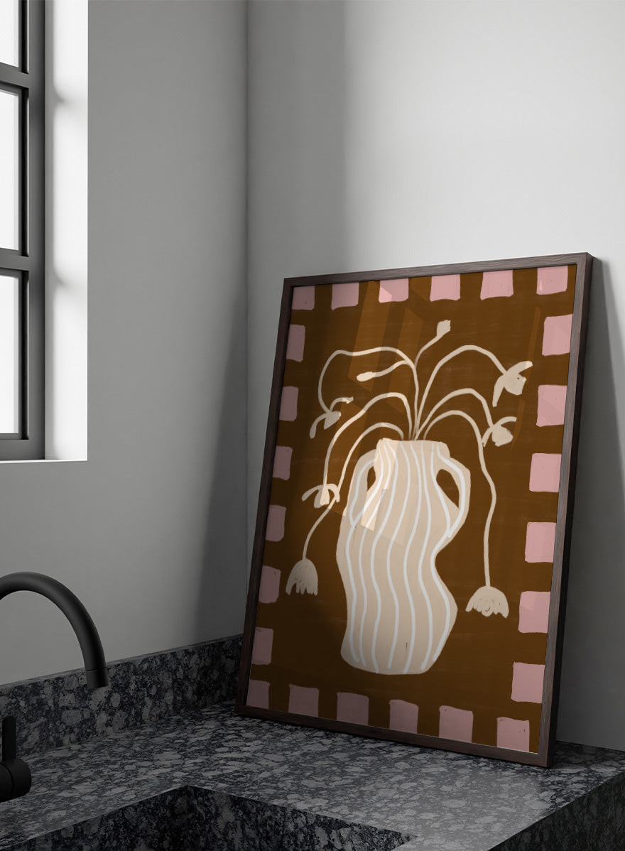 A stylized art print featuring a cream-colored wavy vase with stems and hanging flower buds on a brown background, framed by a pink and brown checkered border. Soft Floral Arrangement by The Art Concept still life art print featuring a striped vase, drooping flowers, warm brown tones, and a soft pink border.