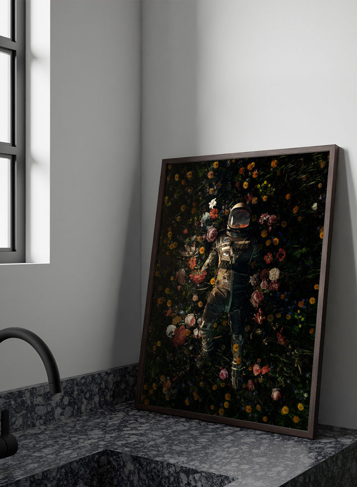 Person in a spacesuit lying on a bed of flowers with a dark background. Cosmic Floral Dream by Nicebleed is a surreal astronaut floral art print featuring a space figure surrounded by flowers, modern aesthetic wall poster.