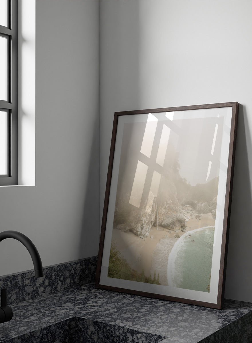 A framed photographic print of a misty coastal beach scene with a small waterfall, sandy shore, and pale blue-green ocean waves. Misty Coastal Haven by Shot by Clint coastal photography print featuring a secluded cove, rocky cliffs, pale turquoise water, and a soft veil of mist.