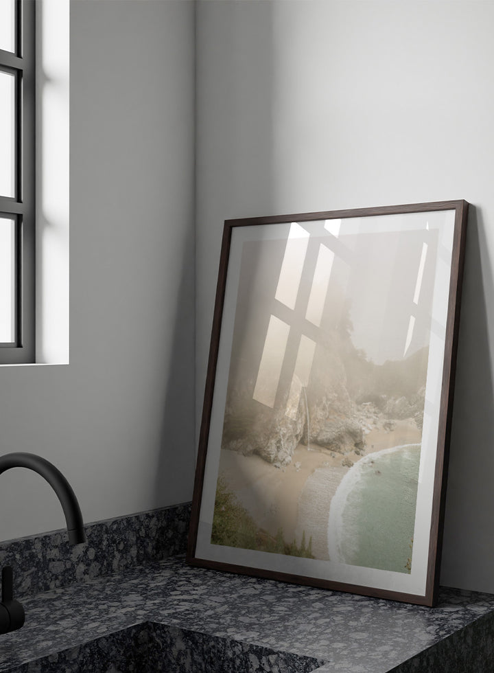 A framed photographic print of a misty coastal beach scene with a small waterfall, sandy shore, and pale blue-green ocean waves. Misty Coastal Haven by Shot by Clint coastal photography print featuring a secluded cove, rocky cliffs, pale turquoise water, and a soft veil of mist.
