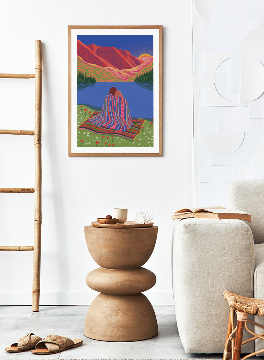 A framed art print featuring a stylized landscape illustration of two people sitting together on a patterned blanket by a blue lake, with red mountains and a yellow sun in the background. Cozy Lake Sunset art print by Ceyda Alasar features a lakeside embrace, vivid mountains, and a glowing sunset in rich, story-filled color.