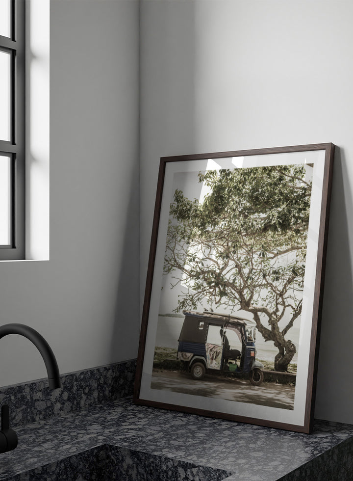 A framed photographic print featuring a small blue and white auto rickshaw parked under a large leafy tree beside a body of water. Tuk Tuk Tour by Shot by Clint travel photography print featuring a blue tuk tuk, surfboard, coastal tree, and relaxed seaside setting.