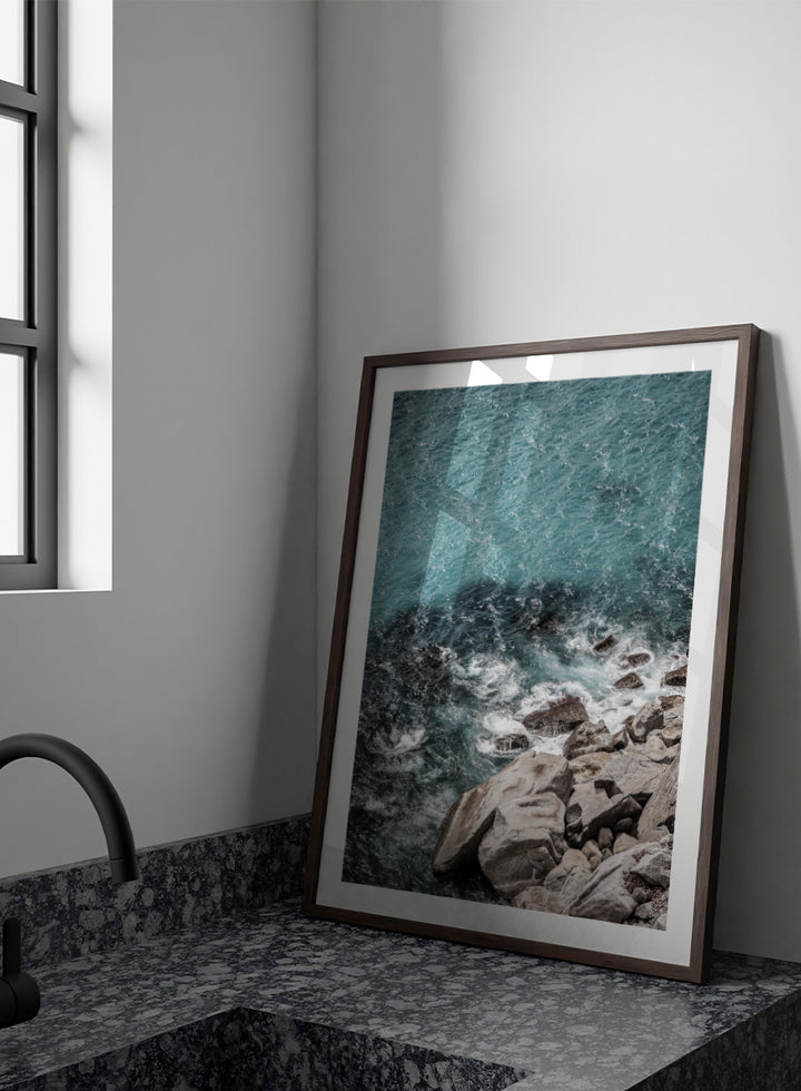 A framed photographic art print depicting a top-down view of turquoise ocean water washing over dark rocks and a rocky shoreline. Ocean’s Edge Symphony by Shot by Clint aerial ocean photography print featuring turquoise water, white surf, coastal texture, and soft seafoam tones.