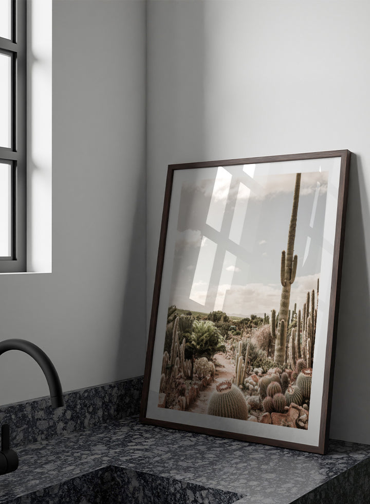 A framed art print depicting a desert landscape with various cacti and a tall saguaro under a cloudy sky. Cactus Farm by Shot by Clint desert photography print featuring towering cacti, winding garden paths, warm stone, and layered botanical texture.