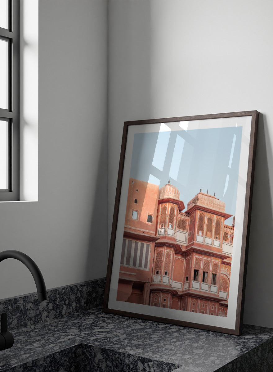 A framed art print featuring a photograph of the iconic pink sandstone Rajasthani palace, Udaipur, with white architectural details under a clear light blue sky. Terracotta Splendor by Shot by Clint architectural photography print featuring ornate palace windows, domed balconies, carved details, and warm coral tones.