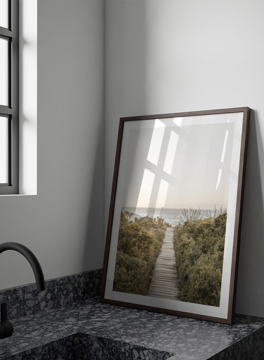 A framed photographic art print depicting a wooden boardwalk leading from dense green beach vegetation to the ocean under a pale sky. Pathway to Tranquility by Shot by Clint coastal art print featuring a wooden boardwalk, wild greenery, distant palms, and a soft ocean horizon.