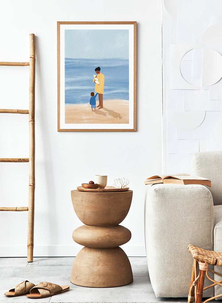 A framed art print featuring an illustration of a man in a yellow striped shirt holding a white dog on a beach next to a child in a blue coat, set against a light blue ocean and pale sky. Family Beach Day by Bea Müller art print featuring a mother with her baby and young child by the sea, created for warm family and coastal decor.