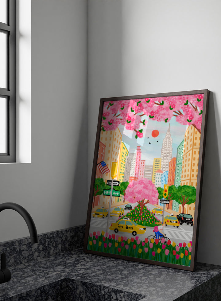 A colorful illustrated city street scene with pink cherry blossoms overhead, yellow NYC taxis, tall buildings, a bicycle with a pink basket, flowers in the foreground, and street signs including Fifti Ave. Shop NYC Cherry Blossoms by Nidhi Kachhadiya, a colorful New York City wall art print with Fifth Avenue, yellow taxis, pink blossoms, and spring city charm.