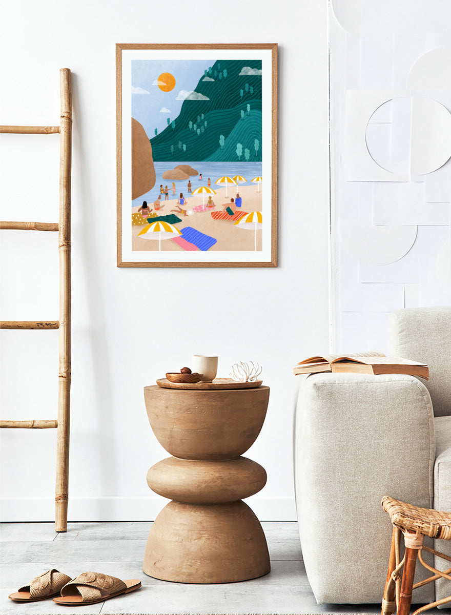 A framed art print featuring a stylized beach scene with a large teal-green mountain, blue water, a yellow sun, and beachgoers under striped umbrellas on a sandy shore. Relaxing Mountain Beach by Bea Müller coastal art print featuring swimmers, striped towels, yellow umbrellas, and a sweeping teal mountain.