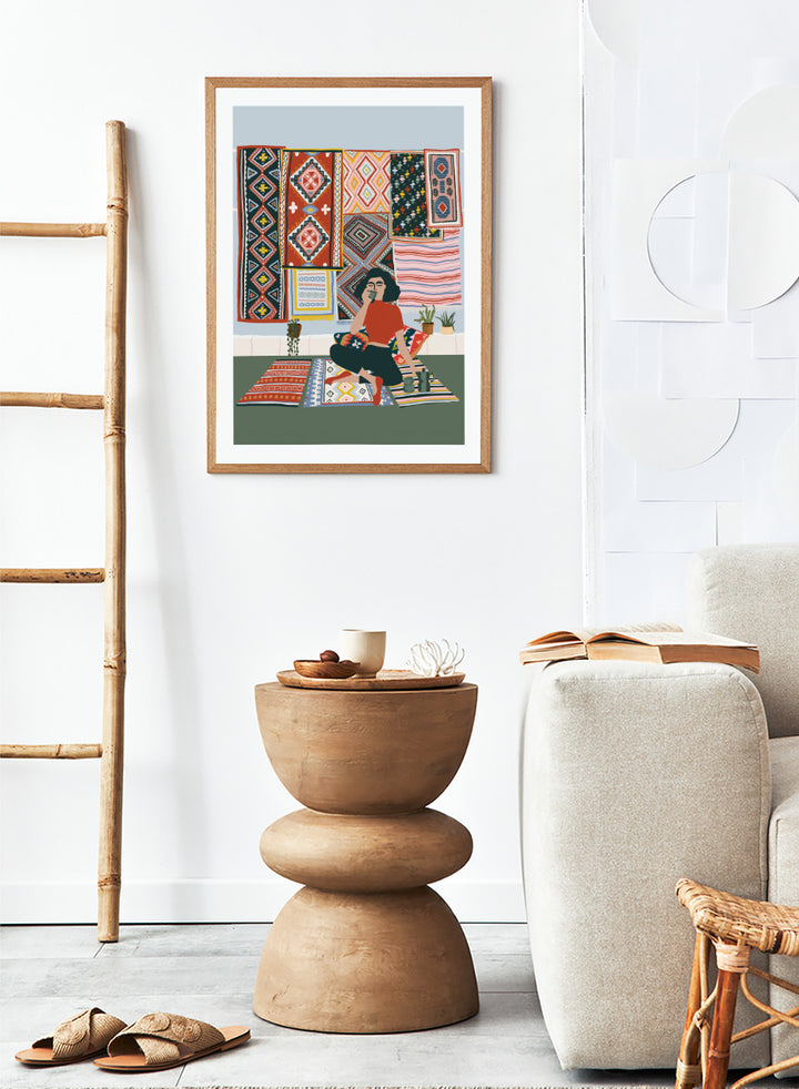 A framed art print featuring a stylized illustration of a woman sitting cross-legged on a patterned rug, surrounded by colorful geometric textiles hanging on a wall. Bohemian Coffee Morning by Ceyda Alasar interior art print featuring patterned rugs, floor cushions, potted plants, coffee, and colorful collected decor.