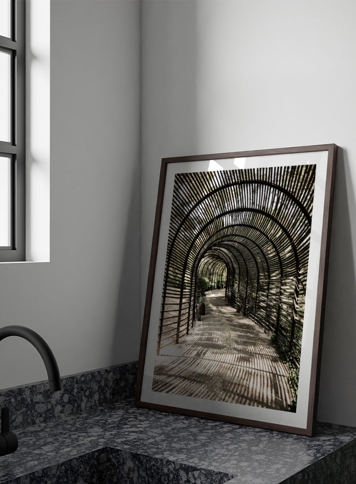 A framed art print featuring a perspective view of a garden pathway enclosed in a dark arched trellis structure, casting striped shadows on the ground. Shadow Garden by Shot by Clint architectural photography print featuring a curved wooden walkway, striped shadows, garden plants, and textured natural light.