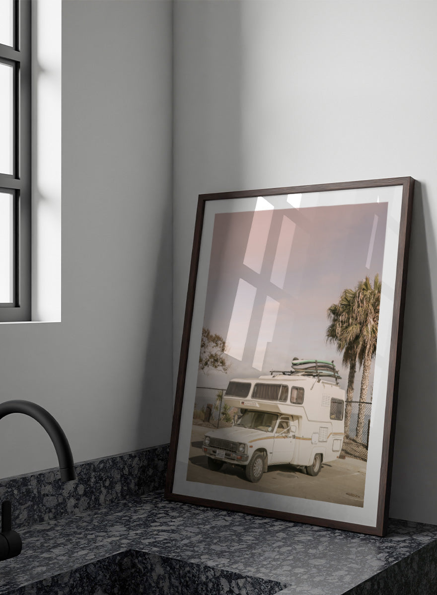 A vintage white camper truck parked outdoors near palm trees and a fence, with a surfboard strapped to its roof rack. Vintage Camper Parked by Shot by Clint travel photography print featuring a retro camper, surfboards, palm trees, and sun-faded coastal tones.