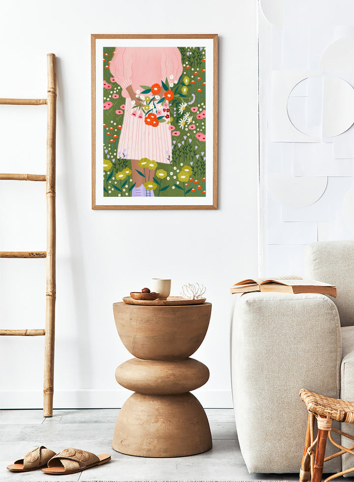 A framed art print featuring a stylized illustration of a person in a pink striped dress holding a bouquet of orange, red, and white flowers in a green field of wildflowers. Secret Garden Bouquet by Bea Müller floral art print featuring a woman gathering wildflowers in a colorful garden filled with poppies, daisies, and greenery.