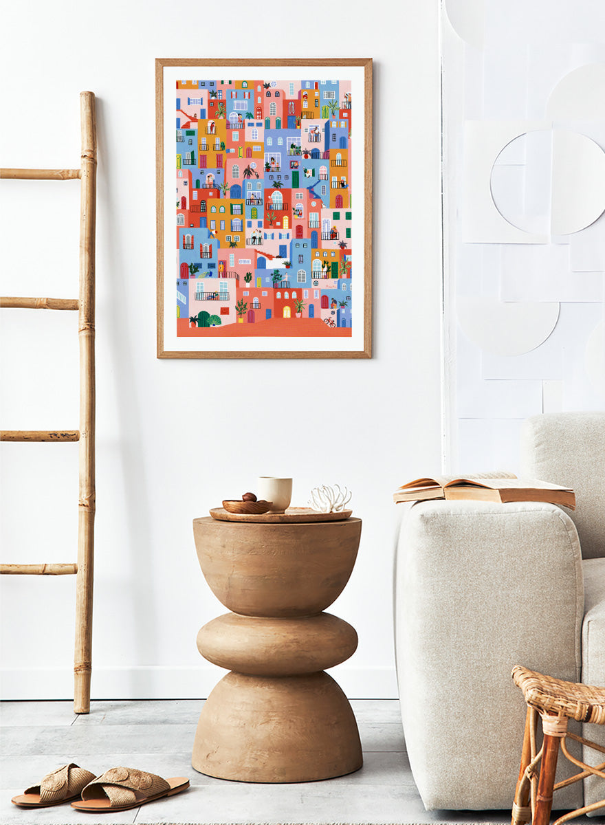 A framed art print featuring a colorful, whimsical illustration of Mediterranean-style buildings with people, animals, and plants. Colorful Town Life by Ceyda Alasar city art print featuring vibrant buildings, balconies, neighbors, plants, and lively urban details in bold color.