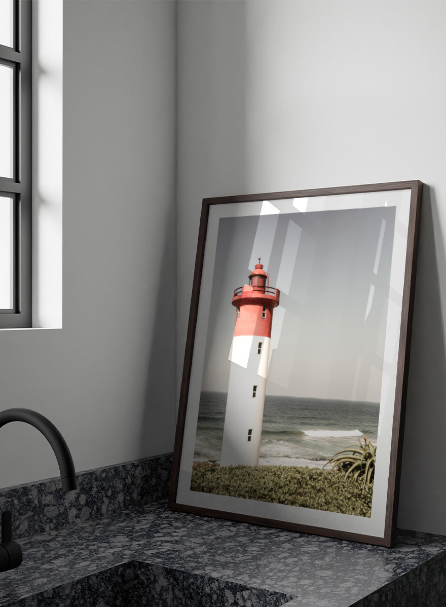 A framed photographic art print of a red and white lighthouse standing on a grassy shore with the ocean and sky in the background. Beacon by the Sea by Shot by Clint coastal photography print featuring a red-and-white lighthouse, ocean waves, pale sky, and coastal greenery.