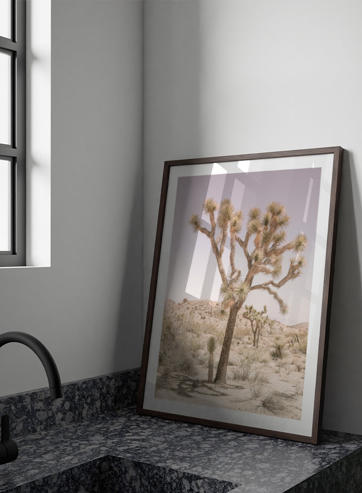 A framed art print depicting a desert landscape with a prominent Joshua tree in the foreground, set against a pale lavender sky and sandy hills. Desert Serenity by Shot by Clint features a Joshua tree desert landscape in soft earthy tones with a pale sky and calm, sun-washed atmosphere.