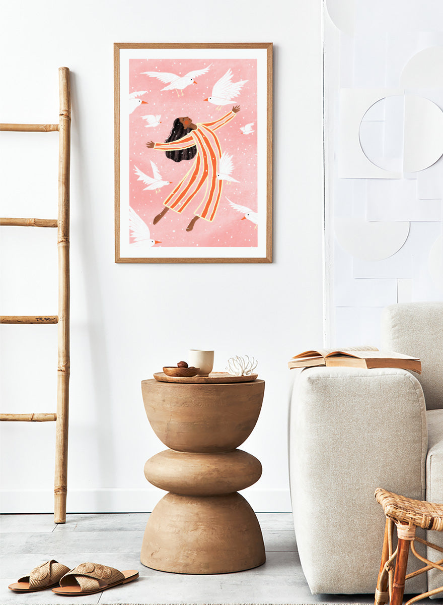 A framed art print featuring an illustration of a woman in orange and white striped pajamas floating among white doves on a soft pink background. Joyful Liberation by Bea Müller woman art print featuring a free-spirited figure surrounded by white birds in bold pink and orange tones.