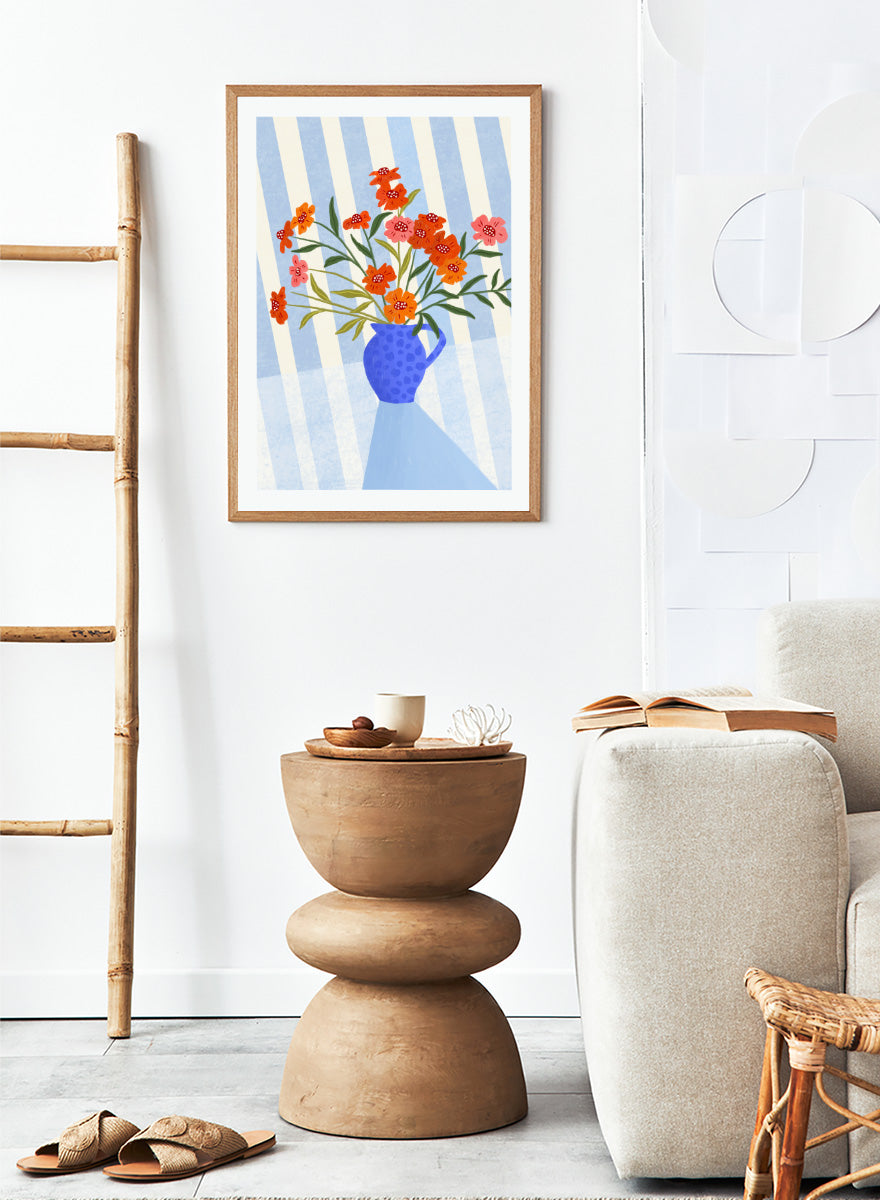 A framed art print featuring a stylized illustration of a blue patterned pitcher vase filled with orange, red, and pink flowers against a blue and white striped background. Cheerful Floral Pitcher by Bea Müller floral still life art print featuring orange and pink flowers in a bright blue jug with striped blue decor.