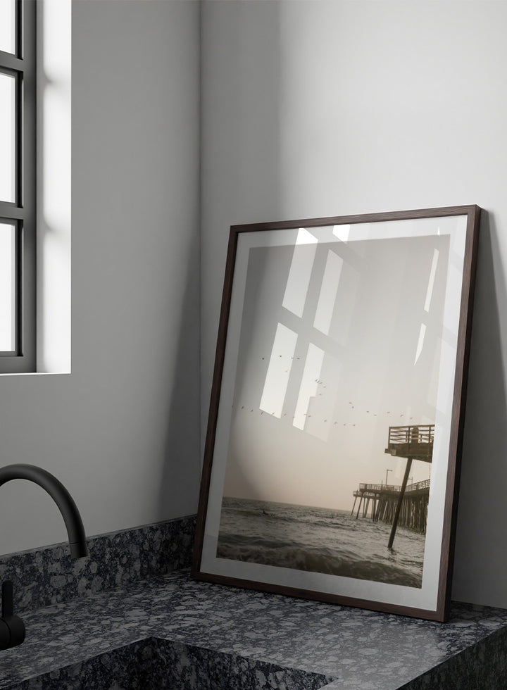 A framed photographic art print depicting a pier extending over a calm ocean under a pale sky, with birds flying across the scene. Seaside Serenity by Shot by Clint coastal photography print featuring ocean waves, a weathered pier, flying birds, and soft sunlit tones.