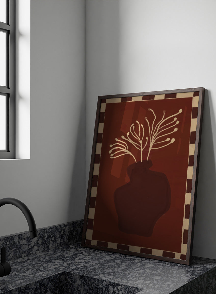An art print featuring a dark brown abstract vase with cream branching stems on a deep reddish-brown background, framed by a beige and dark brown checkered border. Minimalist Vase Plant by The Art Concept still life art print featuring a dark vase, cream stems, checkerboard border, and warm rust-brown tones.