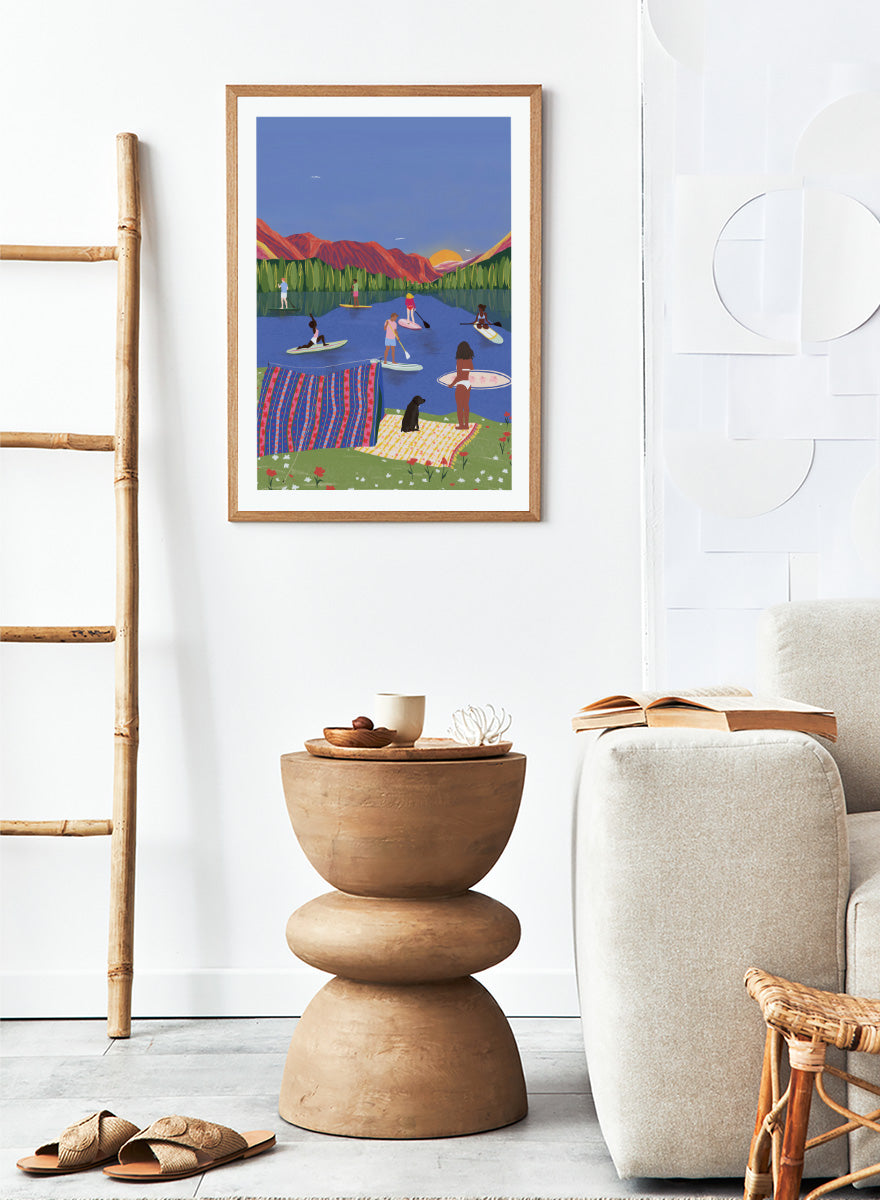A framed art print featuring a colorful, stylized illustration of people paddleboarding and relaxing on a lake with mountains in the background. Paddleboard Lake Life by Ceyda Alasar outdoor art print featuring paddleboarders, mountain scenery, wildflowers, picnic blankets, and a lakeside dog.